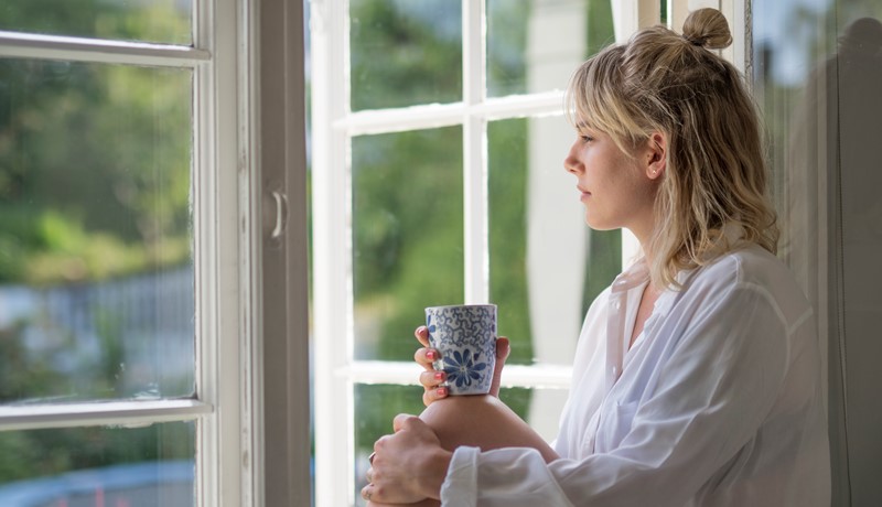 Kvinne med kopp i hånden ser ut av vinduet